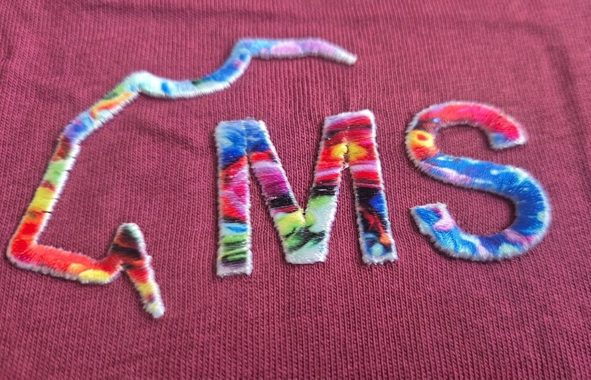 Zwróć uwagę na niesamowicie złożoną mieszankę kolorów wewnątrz liter. Tradycyjna maszyna hafciarska nie ma szans na płynne, fotograficzne przejście tych odcieni przy tak małej powierzchni. Zamiast 'płynności' widzielibyśmy ostre krawędzie nici. Haft 3D z sublimowanym kolorem.